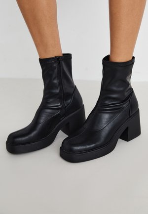 Jambes portant des bottes montantes en cuir noir, de hauteur mi-mollet, à semelle plateforme, talons blocs et fermetures éclair latérales, sur un sol gris clair.