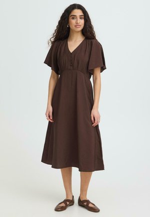 Kvinde med langt krøllet hår iført en brun midi kjole med flagrende ærmer og matchende brune flade sko, stående mod en ensfarvet baggrund.