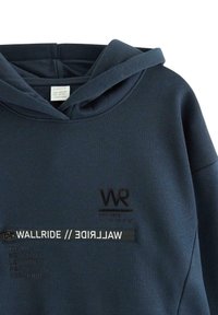 Granatowy hoodie z miękkiego materiału, z logo "WR" z przodu oraz naszywką "WALLRIDE", z nazwami miast wytłoczonymi wzdłuż dolnej krawędzi.