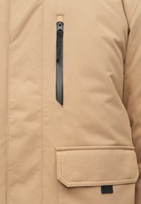 Beige Jacke Nahaufnahme zeigt vertikale schwarze wasserdichte Reißverschlusstasche über großer Klapptasche mit schwarzem Stoff-Zuglasche.