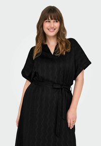 Črna kratka obleka s subtilno zigzag teksturo, V-izrezom in pasom iz blaga. Lahka in tekoča silhueta.