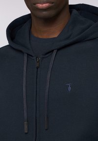 Námořnická modrá mikina s kapucí na zip vyrobená z texturované látky, s přední kapsou, šňůrkovou kapucí a malým vyšitým logem na hrudi.