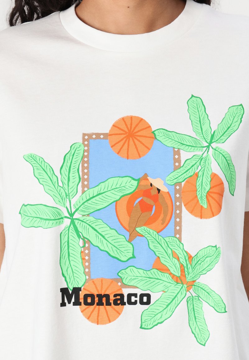T-shirt en coton blanc avec un design graphique coloré comprenant des feuilles vertes, des cercles oranges, un fond bleu et le texte "Monaco" en noir.