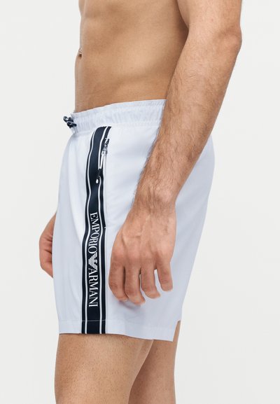 Αρσενικός κορμός και δεξί χέρι που φορούν λευκά μαγιό Emporio Armani με μαύρες κάθετες ρίγες λογότυπου και τσέπη με φερμουάρ στο πλάι.