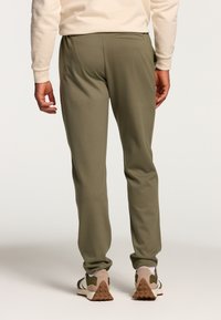 Olive green athletic pants with a tapered fit, elastic waistband, and rear pocket. Material appears smooth and stretchy. Worn with sneakers.