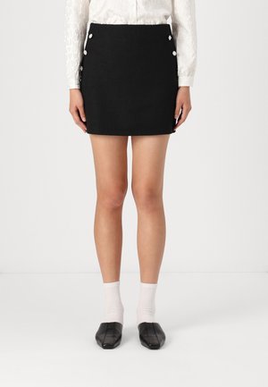 Mini-jupe noire en tissu texturé, avec des accents de boutons argentés sur les côtés. Associée à des chaussures noires et des chaussettes blanches qui arrivent à la cheville.