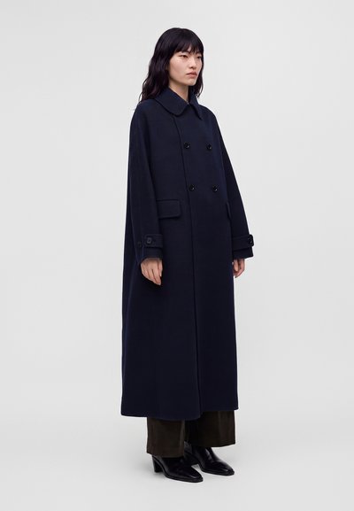 Femme aux longs cheveux noirs portant un long manteau bleu marine foncé à double boutonnage, un pantalon large foncé et des bottes noires à talons, debout devant un fond clair.