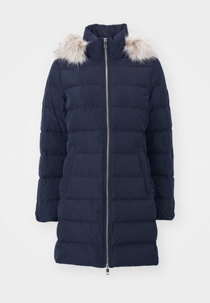 Manteau rembourré bleu marine avec un col montant, fermeture éclair argentée et bordure en fausse fourrure sur la capuche. Comprend un motif matelassé horizontal et des poches latérales.