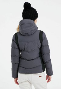Graue Steppjacke mit schwarzen Seitenteilen, Kapuze und gestepptem Design, getragen über weißen Hosen. Ausgestattet mit einer gestrickten schwarzen Mütze mit Pompon.