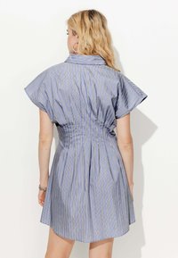 Femme aux cheveux blonds portant une robe à rayures verticales bleues et blanches à manches courtes, debout de dos contre un fond blanc uni.