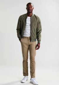 Olivgrüne Bomberjacke über einem weißen gestreiften T-Shirt, kombiniert mit beigefarbenen Slim-Fit-Hosen und weißen Sneakers. Glattes Material und lockere Passform.
