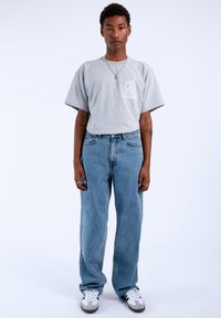 Grijze t-shirt met korte mouwen en een grafisch ontwerp op de borst, gecombineerd met lichtblauwe, loszittende jeans en multikleurige sneakers.