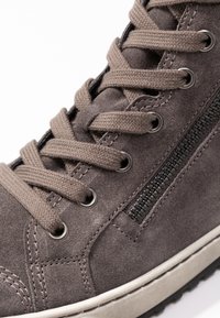 Close-up of a brown suede sneaker with beige laces threaded through metal eyelets and a black side zipper on a white sole.
