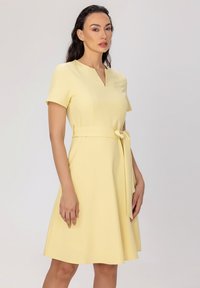 Robe jaune courte avec un décolleté en V, des manches courtes et une ceinture nouée à la taille. Tissu lisse et léger avec une silhouette de jupe évasée.