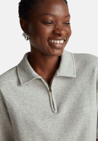 Šedá mikina na zip s límcem, vyrobená z texturovaného materiálu a s předním zipem. Detailní záběr ukazující detaily látky a šití.