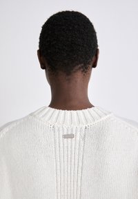 Off-white knit sweater with ribbed collar and hem. Features a metal label at the back. Texture appears soft with a loose weave.