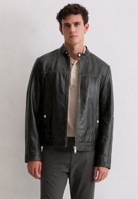 Mann, der eine schwarze Lederjacke mit Reißverschluss über einem beigen Hemd mit Knöpfen und dunklen Hosen trägt, steht vor einer schlichten weißen Wand.