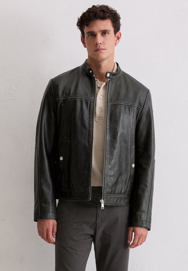 Mann, der eine schwarze Lederjacke mit Reißverschluss über einem beigen Hemd mit Knöpfen und dunklen Hosen trägt, steht vor einer schlichten weißen Wand.