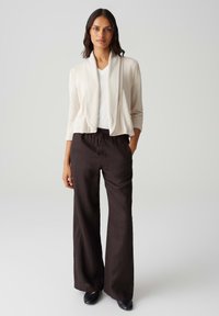 Hellbeige cardigan mit offenem Front, drei Viertelärmel, über einem weißen V-Ausschnitt-Shirt, kombiniert mit lockeren, dunkelbraunen weitem Hosen.