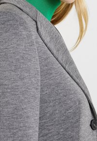 Felpa grigia con texture liscia, maniche raglan e bottoni neri sul davanti. Accentata da una maglia verde a collo alto sotto.