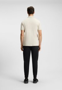 Lichte beige polo met korte mouwen voor heren, gecombineerd met zwarte joggerbroeken en zwarte sportschoenen, gezien vanuit de achterkant.