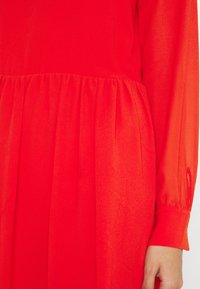 Punainen mekko, jossa on kerätty vyötärö, pitkät läpinäkyvät hihat ja sileä, kevyt kangas. Väri on kirkas ja tasainen koko mekossa.