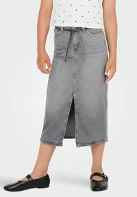 Pilkos džinsinės midi sijonas su priekinėmis skeltinėmis, aukštu liemeniu ir kaklaraiščio diržu. Dėvimas su juodomis plokščiomis batais, pasižyminčiais lygiu tekstūru ir kasdiene išvaizda.