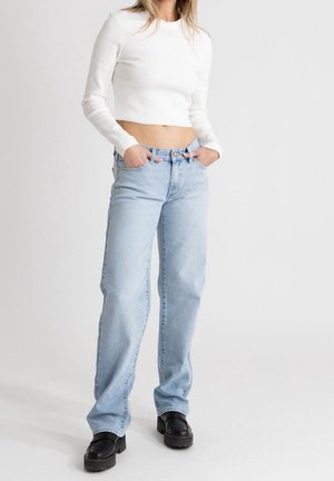 Osoba ubrana w biały, długi krótki top, jasnoniebieskie, wysokie dżinsy oraz czarne buty na platformie, z rękami w kieszeniach.