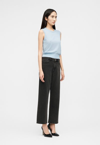 Frau steht, trägt ärmelloses hellblaues Oberteil, hoch taillierte lockere schwarze Jeans und schwarze High-Heels, vor schlichtem weißem Hintergrund.