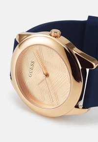 Montre en or rose avec un cadran texturé présentant des motifs géométriques, montée sur un bracelet en silicone bleu. Comprend une couronne latérale pour le réglage.