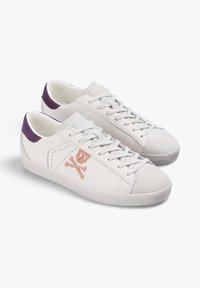 Zapatillas de cuero blancas con un talón de ante morado, que cuentan con un diseño de calavera y huesos cruzados en rosa y una suela de goma blanca texturizada.