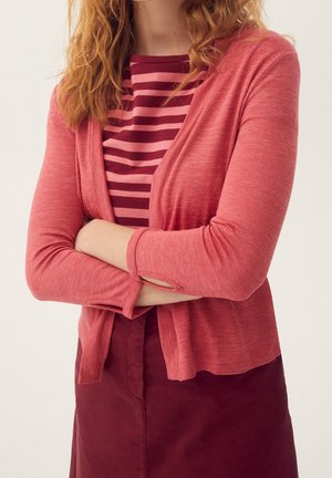 Lichtroze cardigan met lange mouwen, open voorkant en opgerolde manchetten, gecombineerd met een gestreept topje. Het materiaal lijkt zacht en textuurrijk.