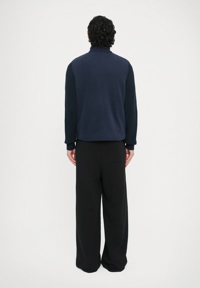 Jachetă fleece bleumarin cu guler înalt și mâneci striate, asortată cu pantaloni negri cu picioare largi și buzunar lateral.