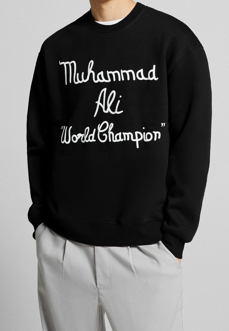 Personne portant un sweat-shirt noir avec un texte blanc indiquant "Muhammad Ali 'World Champion'", associé à un pantalon gris clair.