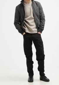 Person som bär en svart jacka, ljusbeige sweatshirt, svarta byxor med dragkedjefickor och svarta sneakers, står med händerna i fickorna.