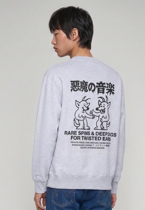 Ung mand set fra siden iført lysegrå sweatshirt med sorte tegneserie-dæmoner og tekst om musik på ryggen.