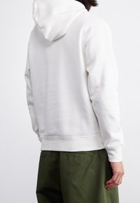 Felpa bianca con un tessuto morbido e strutturato, dotata di cappuccio posteriore e vita aderente. Abbinata a pantaloni verde scuro, larghi e comodi.