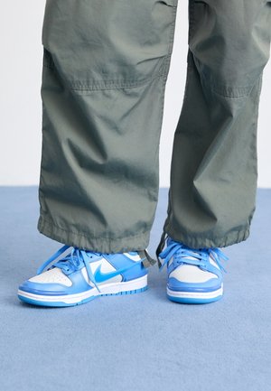 Niebiesko-białe sneakersy Nike noszone z luźnymi oliwkowozielonymi spodniami, stojące na jasnoniebieskiej podłodze.