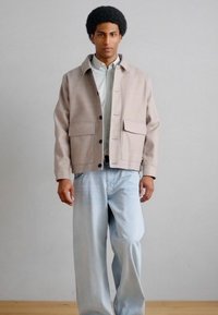 Άνδρας που φοράει μπεζ σακάκι πάνω από ριγέ πουκάμισο και ανοιχτό μπλε τζιν παντελόνι με φαρδιά μπατζάκια, στέκεται μπροστά σε ουδέτερο φόντο.