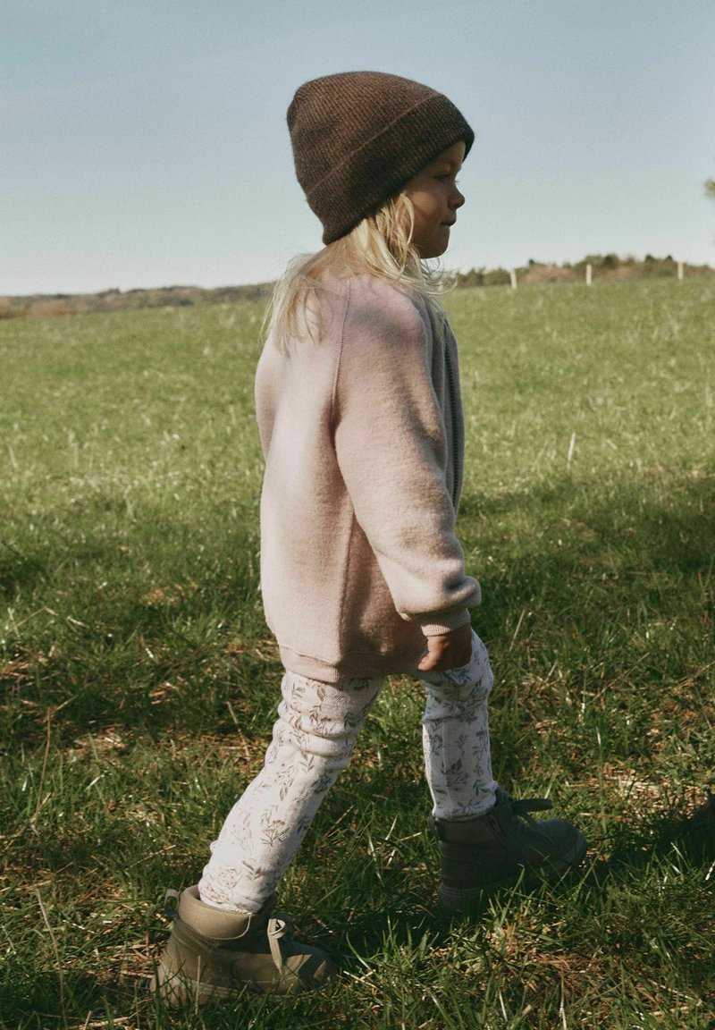 Jeune enfant portant un bonnet tricoté marron, un pull beige surdimensionné, des leggings fleuris et des bottes, marchant sur l'herbe dans un champ ouvert sous un ciel dégagé.