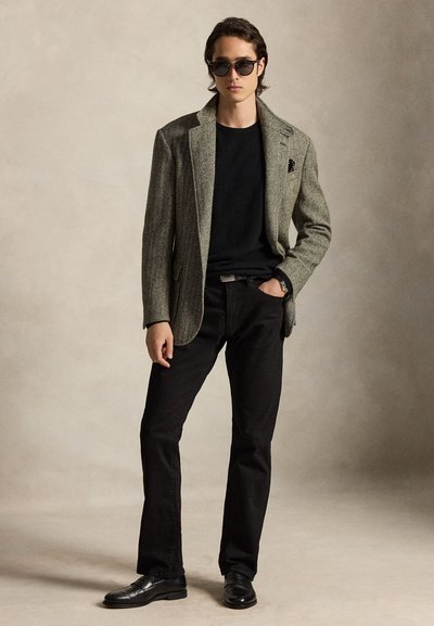 Junger Mann mit grauem Blazer, schwarzem Hemd, schwarzer Hose, schwarzen Schuhen und Sonnenbrille, steht mit einer Hand in der Tasche vor neutralem Hintergrund.