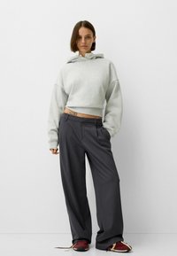 Felpa grigia corta con cappuccio e maniche oversize abbinata a pantaloni larghi grigio scuro e scarpe da ginnastica bordeaux con lacci a contrasto.