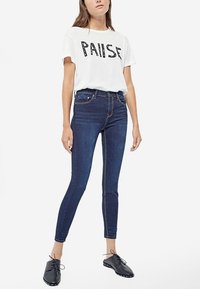 Camiseta blanca con la palabra "PAISE" en letras negras, combinada con jeans ajustados de color azul oscuro y zapatos negros con cordones, presentando un diseño entallado.