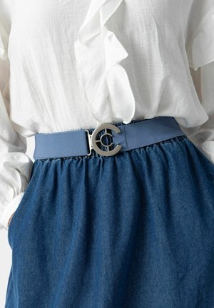 Curea din material textil albastru cu o cataramă circulară argintie. Curea are o textură netedă și este purtată peste o fustă din denim și o bluză albă cu volane.