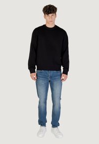 Joven de pie, vistiendo una sudadera negra, jeans azules y zapatillas blancas contra un fondo claro y liso.