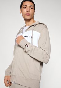 Μπεζ φούτερ με φερμουάρ και λευκή πάνελ, τύπωμα με το κείμενο "Champion". Ριμπ μανσέτες, ρυθμιζόμενο σκουφάκι με κορδόνι, πλαϊνές τσέπες και λογότυπο σε μπαλώμα.