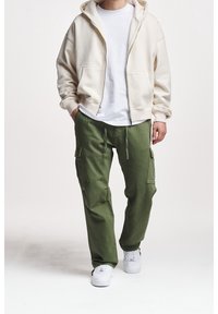 Felpa beige con zip e una consistenza morbida, indossata sopra una t-shirt bianca. Pantaloni cargo verde oliva con tasche e una vestibilità rilassata, abbinati a sneaker bianche.