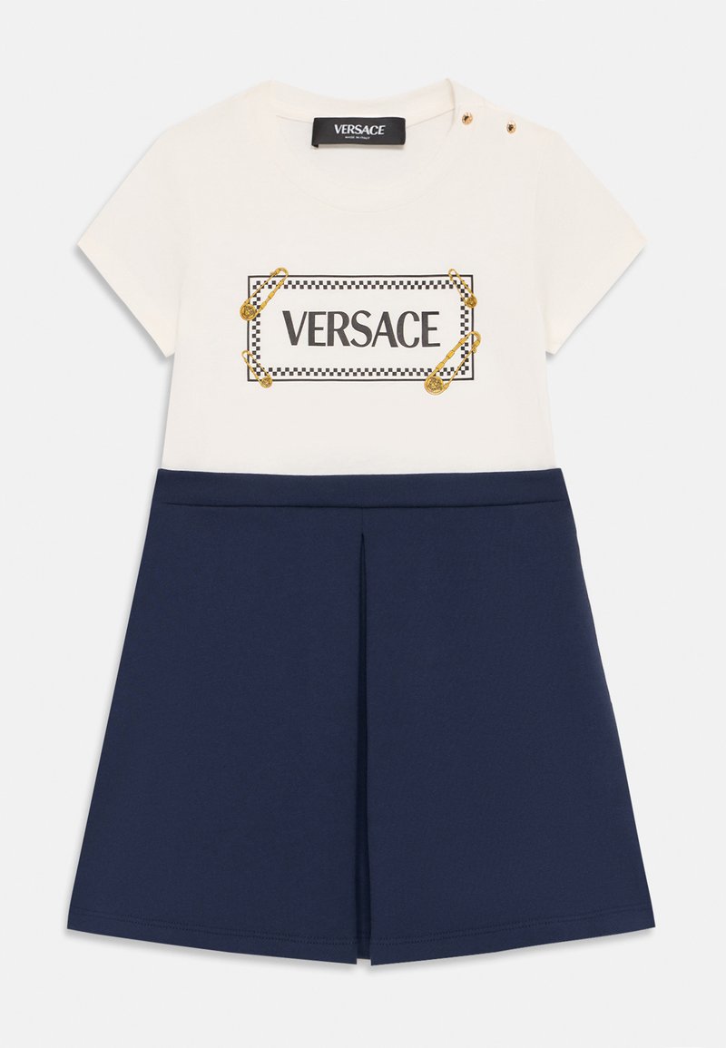 T-shirt de algodón blanco con el logo "VERSACE" y detalles de alfileres de seguridad dorados, acompañado de una falda en línea A de color azul marino oscuro con un detalle de pliegue.