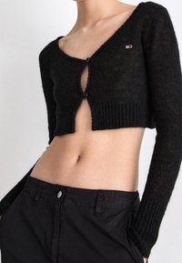 Suéter negro cropped con mangas largas, textura acanalada y un escote profundo, que presenta dos cierres de botón en la parte frontal.