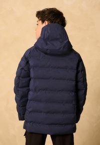 Personne avec les cheveux courts et foncés portant une veste matelassée bleu marine avec capuche, de dos contre un fond beige.